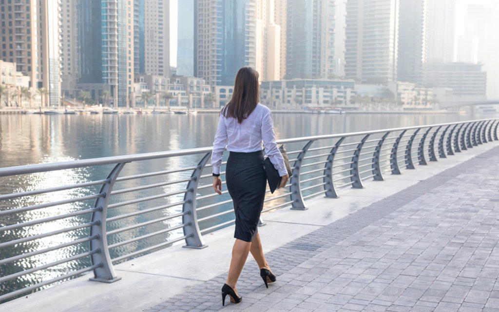Mulher de negócios caminha por uma ponte em Dubai com um lago e os prédios modernos ao fundo