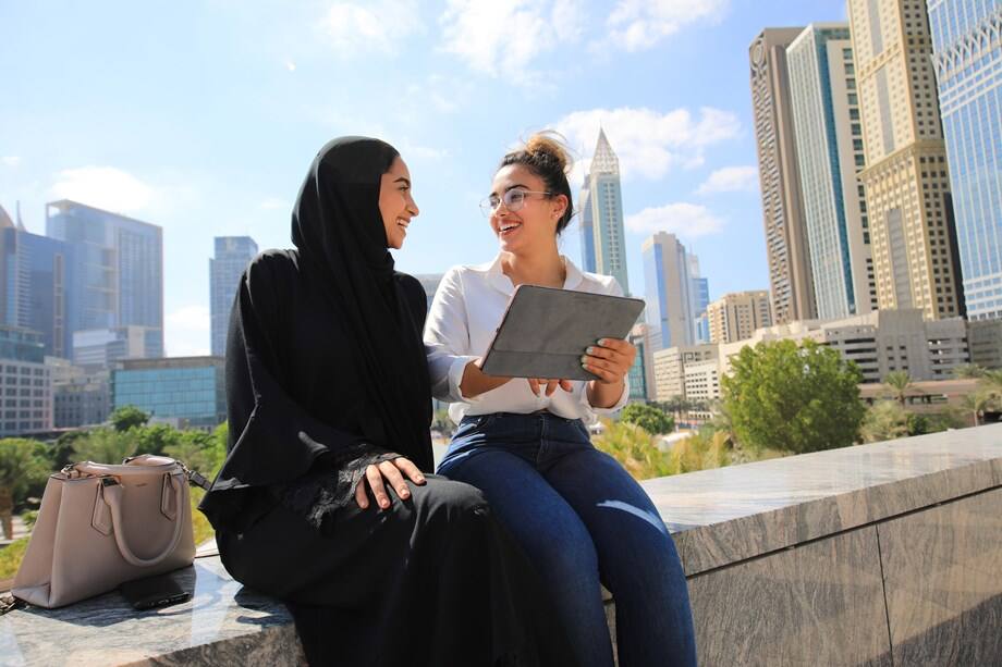 Duas mulheres, uma de burca e outra sem, simbolizando estudantes em Dubai conversando enquanto uma delas segura um tablet