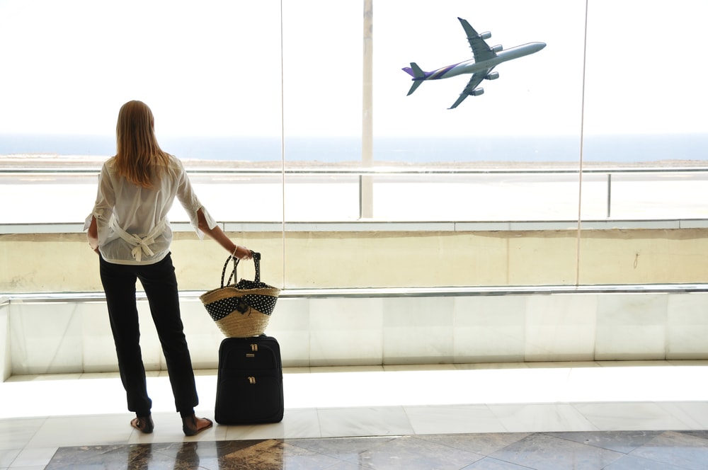 Mulher olhando um avião no céu pela janela do aeroporto segurando uma mala | Planejamento de intercâmbio